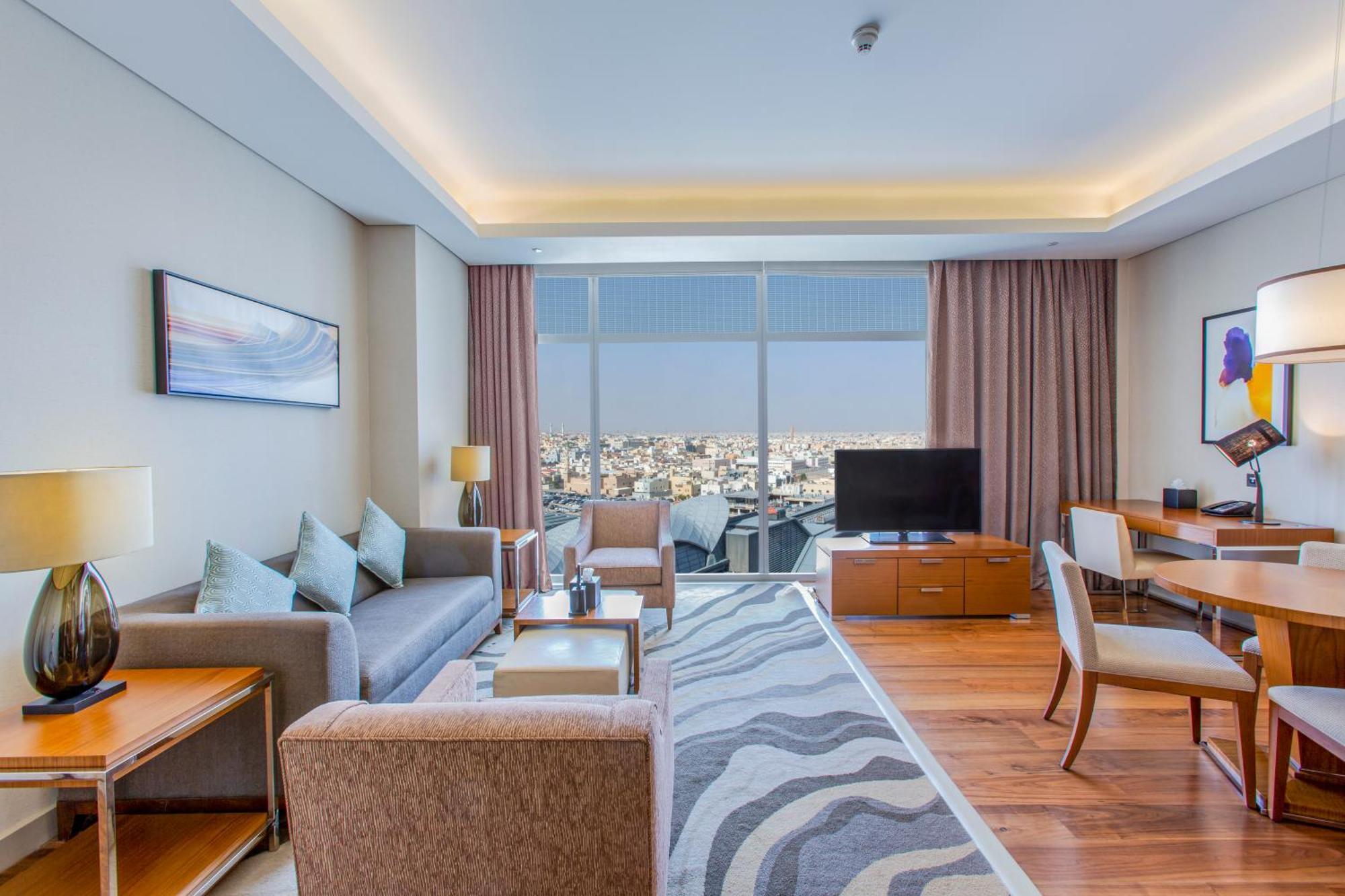 Hilton Riyadh Hotel & Residences Εξωτερικό φωτογραφία The photo shows a modern hotel room or suite. The space features a stylish living area with a sofa and armchairs arranged around a coffee table. There are lamps providing ambient lighting, and a large window offers a view of the cityscape outside. A 