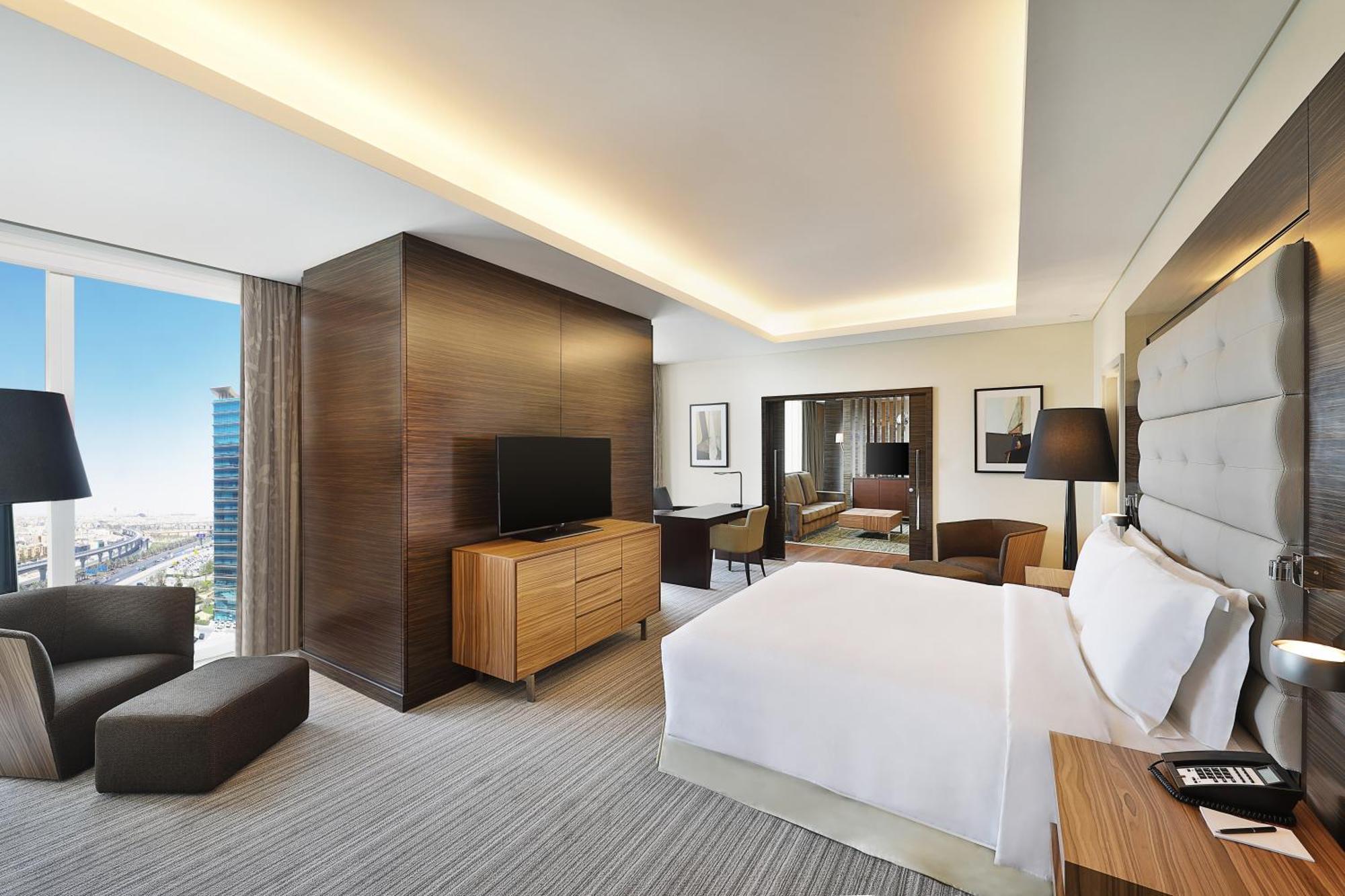 Hilton Riyadh Hotel & Residences Εξωτερικό φωτογραφία The photo shows a modern hotel room. It features a neatly made bed with white linens, a wooden nightstand beside it, and a cozy armchair. The room has a large window that provides a view of the outside, likely showcasing a cityscape. A flat-screen TV