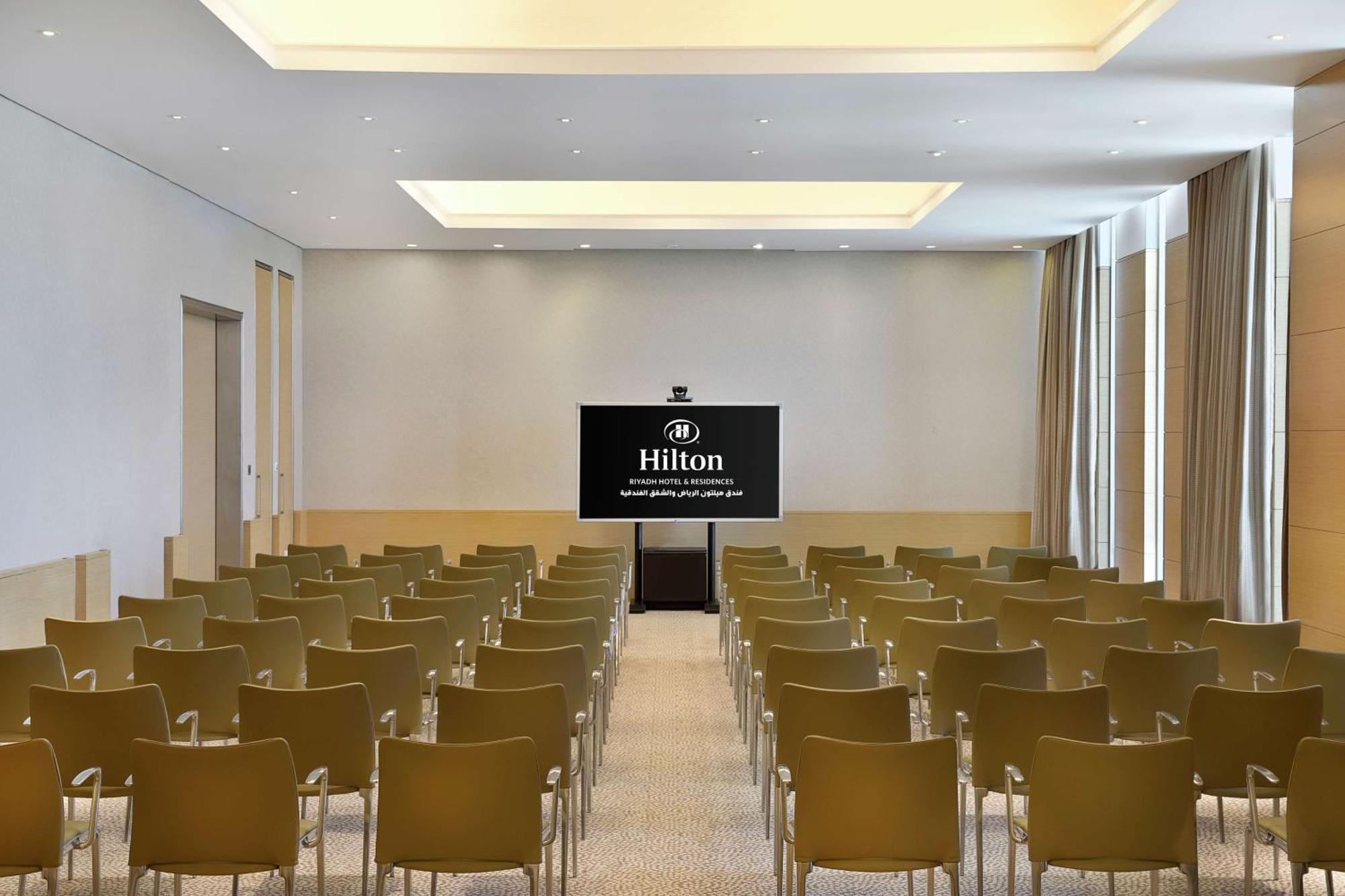 Hilton Riyadh Hotel & Residences Εξωτερικό φωτογραφία The photo shows a conference room or meeting space. It features rows of green chairs arranged facing a large screen that displays the Hilton logo. The room has a modern design with light-colored walls and a bright ceiling. Natural light appears to co