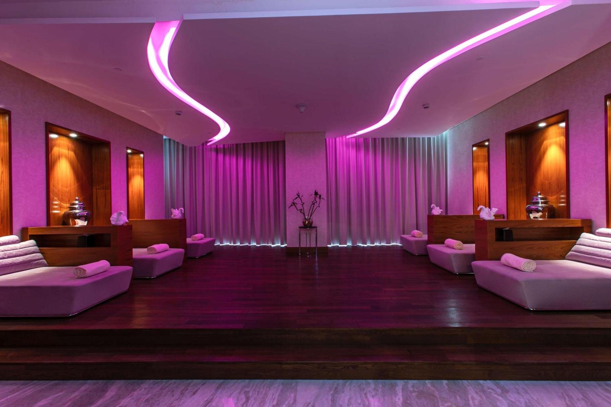 Hilton Riyadh Hotel & Residences Εξωτερικό φωτογραφία The photo depicts a relaxing interior space, likely a spa or wellness area. It features a wooden floor with a raised platform accented by soft, low couches on either side. The walls are adorned with modern lighting, which emits a calming pink hue. Th