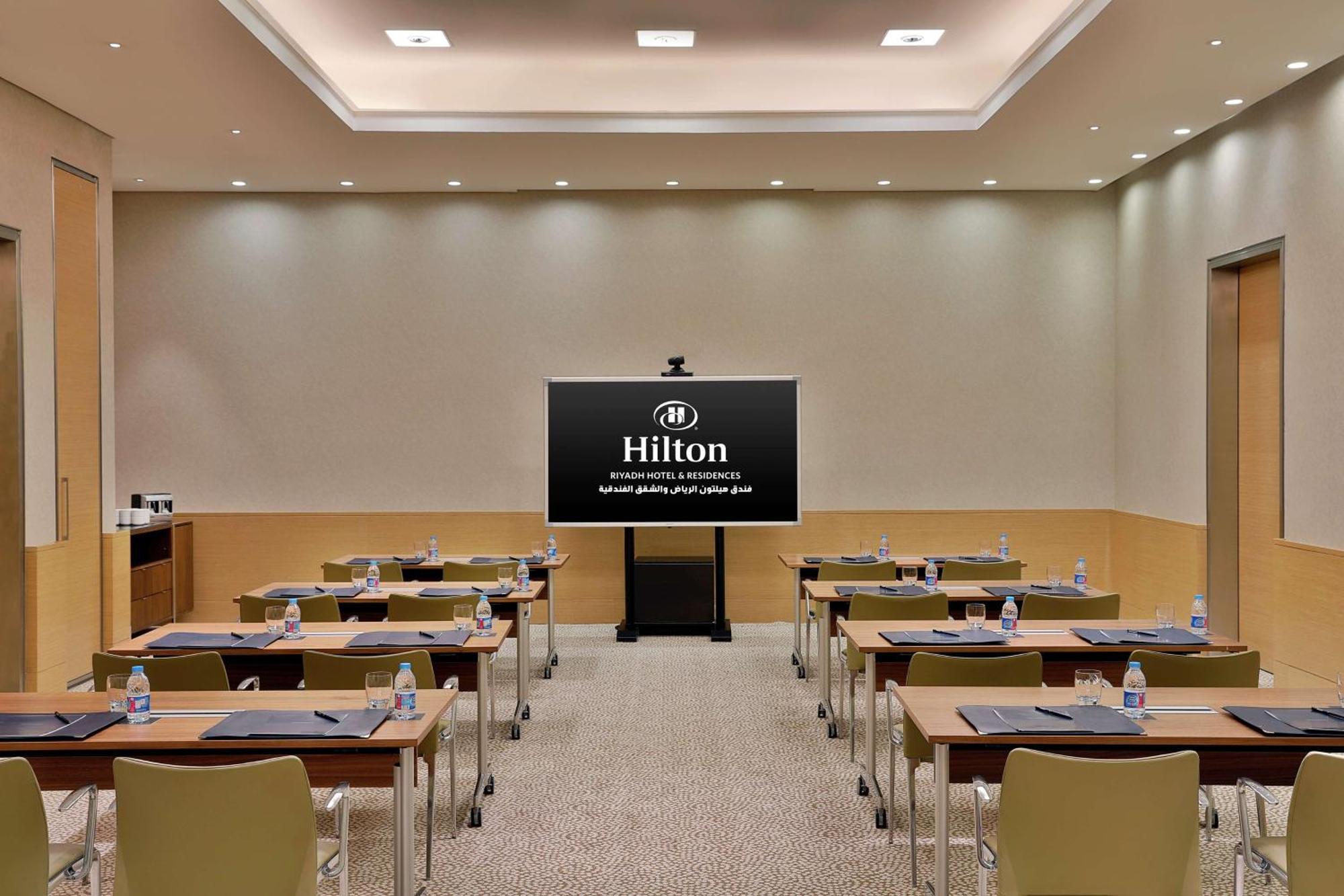 Hilton Riyadh Hotel & Residences Εξωτερικό φωτογραφία The photo shows a conference or meeting room set up for a presentation. There are several tables arranged in a classroom style, each equipped with chairs. On each table, there are notepads and water bottles. At the front of the room is a large screen