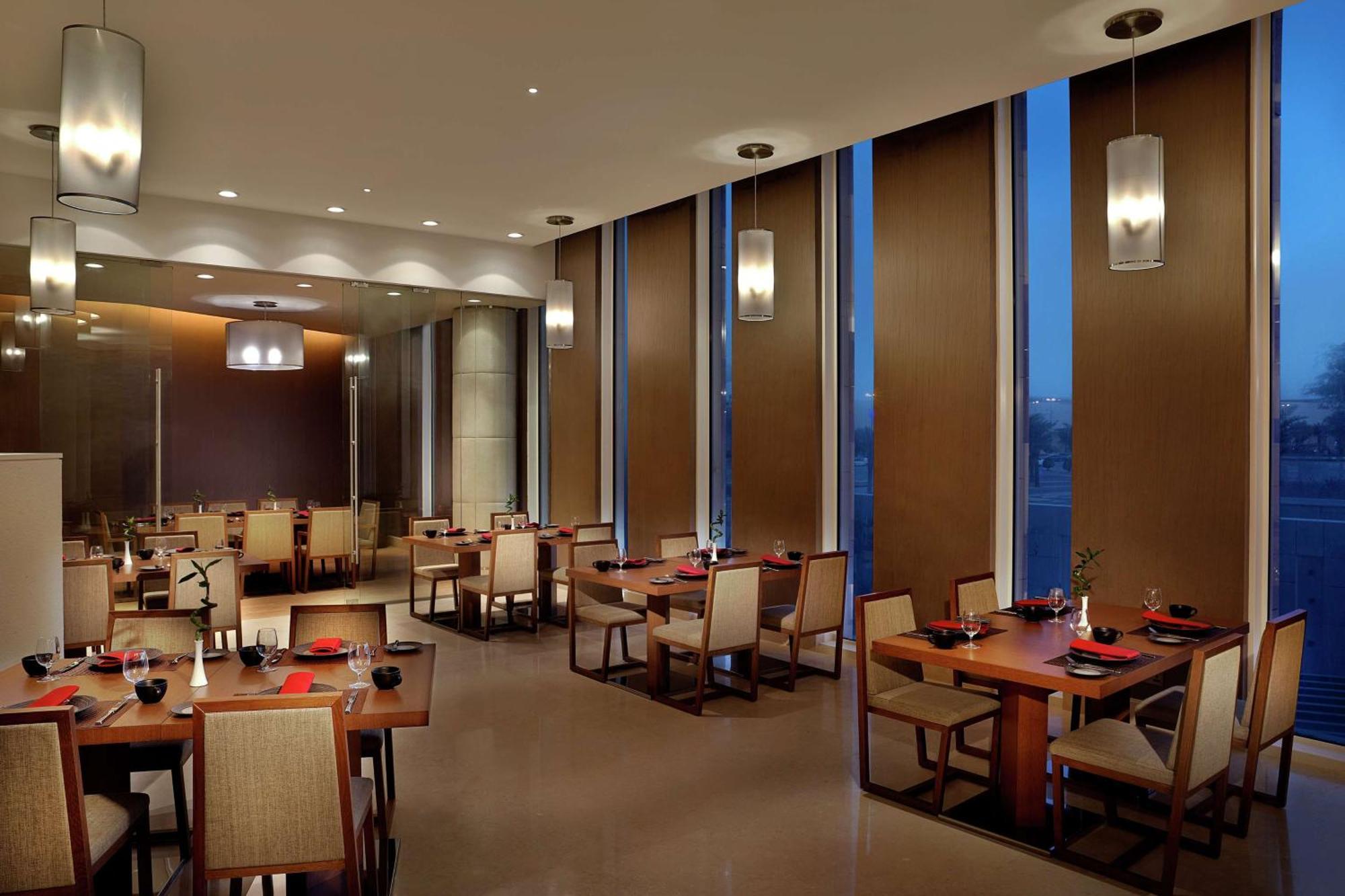 Hilton Riyadh Hotel & Residences Εξωτερικό φωτογραφία The image depicts a modern restaurant interior. It features several dining tables arranged neatly, each set with plates and utensils. The room has large windows with blinds partially drawn, allowing some ambient light to filter in, while evening ligh