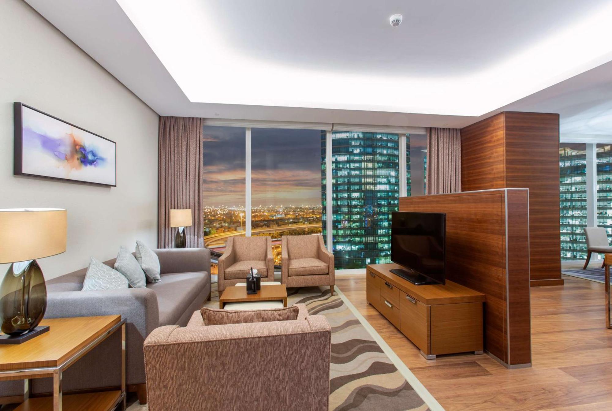 Hilton Riyadh Hotel & Residences Εξωτερικό φωτογραφία The photo shows a modern living room in a stylish apartment. The space features a comfortable seating arrangement with a light gray sofa and two brown armchairs, complemented by a wooden coffee table. There is a large flat-screen TV mounted on a wood