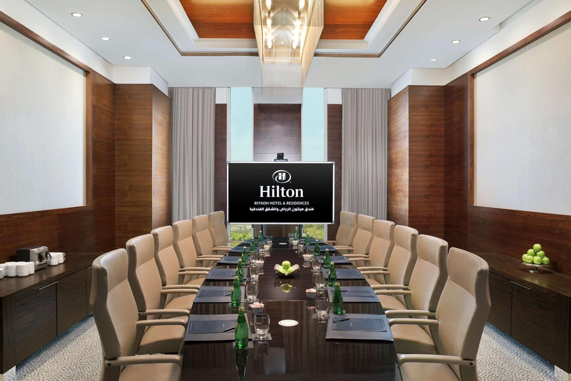 Hilton Riyadh Hotel & Residences Εξωτερικό φωτογραφία The photo shows a modern conference room within a Hilton hotel. There is a large wooden table surrounded by several comfortable chairs. At the head of the table is a large screen displaying the Hilton logo. The room is well-lit with stylish overhead 