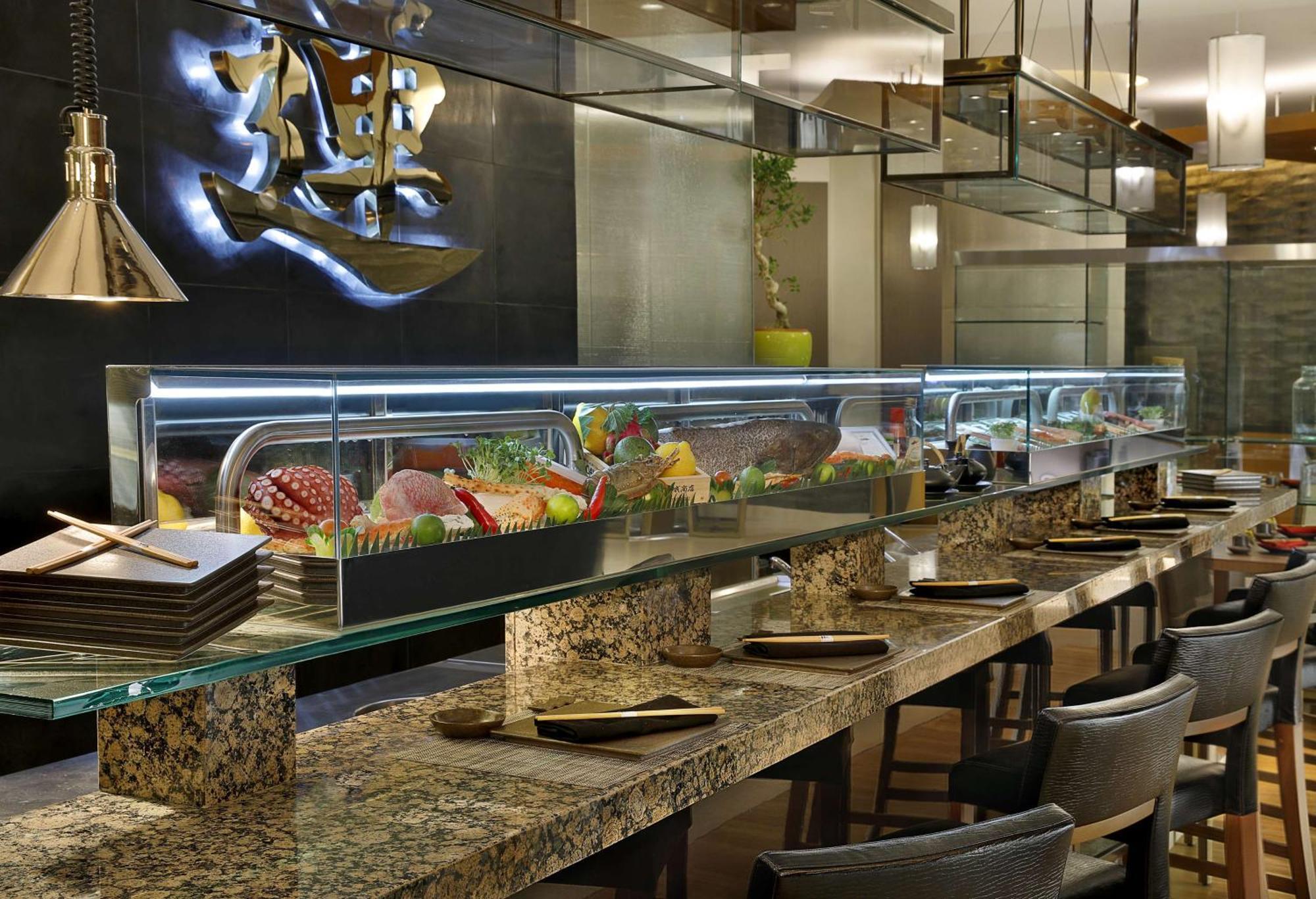 Hilton Riyadh Hotel & Residences Εξωτερικό φωτογραφία The photo shows an interior view of a sushi restaurant. There is a long, sleek sushi bar with a glass display case filled with various sushi and sashimi offerings. The counter is made of polished stone, and there are several dark chairs arranged neat